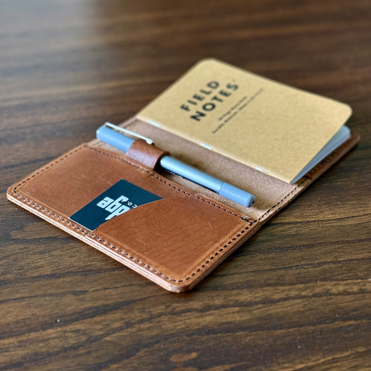 Brown leather notebook cover with 'Field Notes' notebook and blue pen on a wooden surface